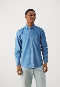 Camisa de mezclilla azul con botones, de manga larga, con un bolsillo en el pecho y cuello. Combinada con jeans azul claro. Fondo plano.