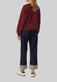 Donna che indossa un top a maniche lunghe bordeaux, jeans larghi scuri con risvolto e scarpe da ginnastica bianche, in piedi davanti a uno sfondo semplice.