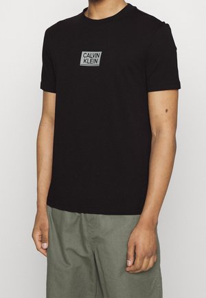Homme portant un t-shirt noir Calvin Klein à manches courtes et un pantalon vert olive, debout devant un fond blanc uni.