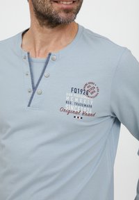 Világoskék, hosszú ujjú pamut keverék ing. Jellemzője egy henley nyak kivágás, három gombbal, navy kiegészítőkkel és különböző színekben nyomtatott szöveggel.