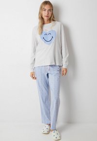 Grijs shirt met lange mouwen en een blauwe hart- en smiley-gezichtafdruk, gecombineerd met lichtblauwe gestreepte broek met kleine blauwe stippen. Witte slippers met blauwe harten.