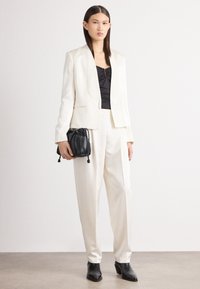 Blazer branco ajustado e calças combinando com uma blusa preta justa, segurando uma pequena mala preta com cordão. Texturas suaves e linhas limpas.