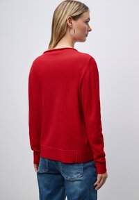 Maglione rosso con orlo e polsini a coste, dotato di scollo rotondo. Il tessuto sembra morbido con una consistenza liscia. Indossato su jeans blu.