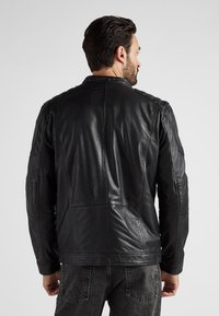 Mann mit dunklen Haaren, der eine enganliegende schwarze Lederjacke und schwarze Jeans trägt, mit dem Rücken zu einem schlichten hellen Hintergrund gewandt.