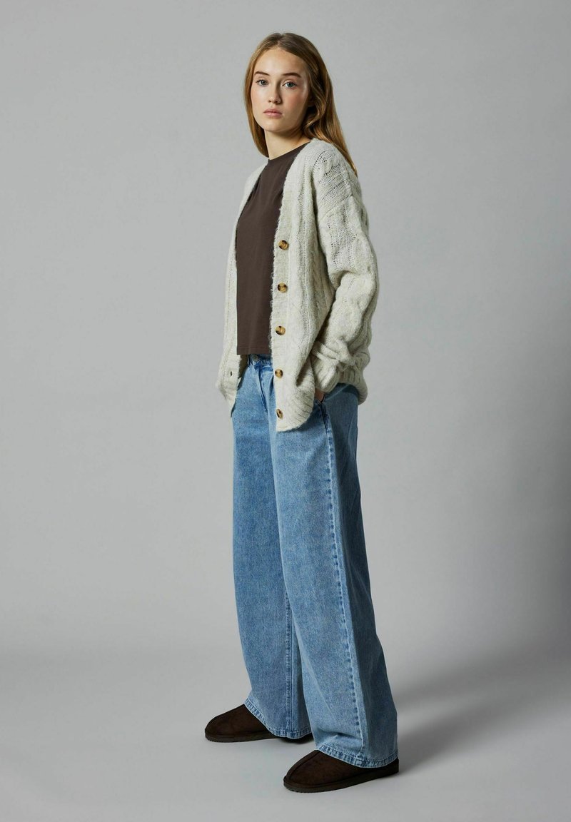 Cardigan lavorato a maglia grigio chiaro con bottoni di legno, indossato sopra una maglia marrone scuro, abbinato a jeans blu a gamba larga e calzature marrone scuro.