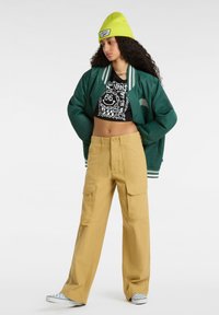 Giacca bomber verde con strisce bianche, crop top grafico nero, pantaloni cargo gialli a gamba larga e un berretto verde brillante. Sneakers blu con motivi.