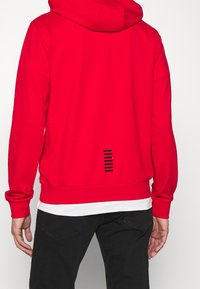 Rote Kapuzenjacke mit glatter Textur, ausgestattet mit einem schwarzen Streifen-Detail am unteren Rücken sowie gerippten Bündchen und Saum.
