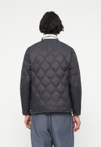 Quilted mørkegrå jakke med rund hals og lange ermer, som har et strukturert mønster og trykknaplukking ved hemmet.