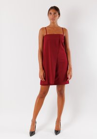 Abito A-line borgogna con spalline sottili, caratterizzato da un drappeggio laterale. Tessuto liscio con una leggera lucentezza, abbinato a scarpe con tacco alto nere.