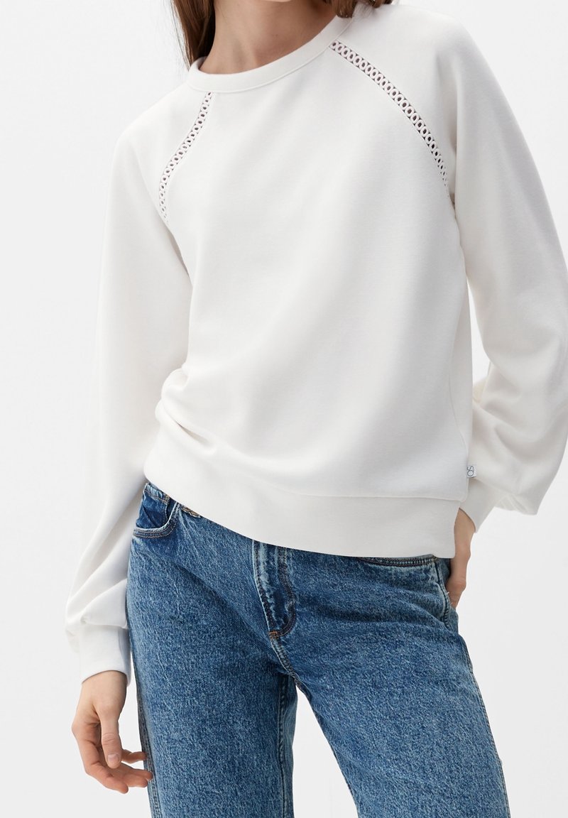 White sweatshirt with raglan sleeves, featuring perforated detailing along the seams. Paired with blue jeans, showcasing a relaxed fit.