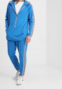 Chándal azul que incluye una sudadera con cremallera y bolsillo tipo canguro, junto con pantalones a juego. Adornado con franjas laterales con patrón y el logo de Nike.