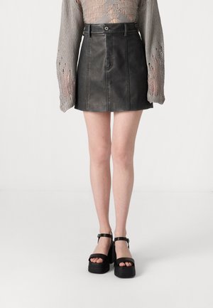 Schwarzer Minirock aus Leder mit sichtbaren Nähten. Kombiniert mit klobigen schwarzen Plateausandalen mit breiten Riemen und einem Knöchelverschluss.