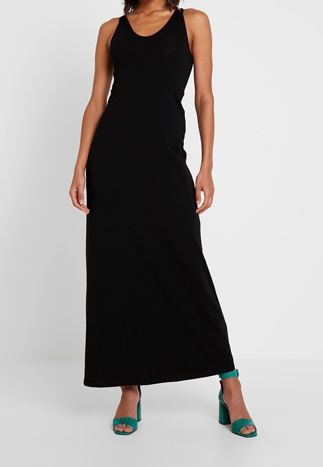 Femme portant une robe maxi noire sans manches associée à des sandales à talons blocs ouvertes teal, se tenant devant un fond blanc uni.