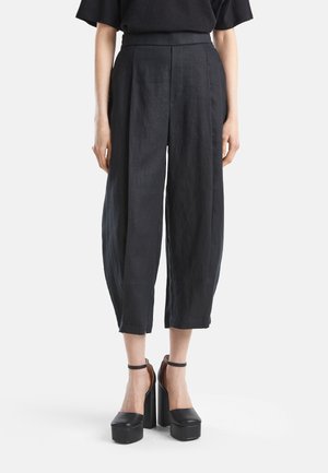 Vrouw draagt zwarte wijde cropped broek met hoge taille en zwarte plateauhakken met enkelbandjes, staand tegen een witte achtergrond.