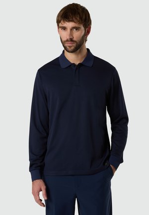 Uomo con barba che indossa una polo a maniche lunghe blu scuro e pantaloni abbinati, in piedi davanti a uno sfondo chiaro e uniforme.
