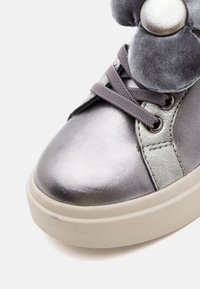 Zapatilla metálica plateada con suela blanca gruesa, cordones grises y una decoración de felpa en forma de pata cerca de los cordones.