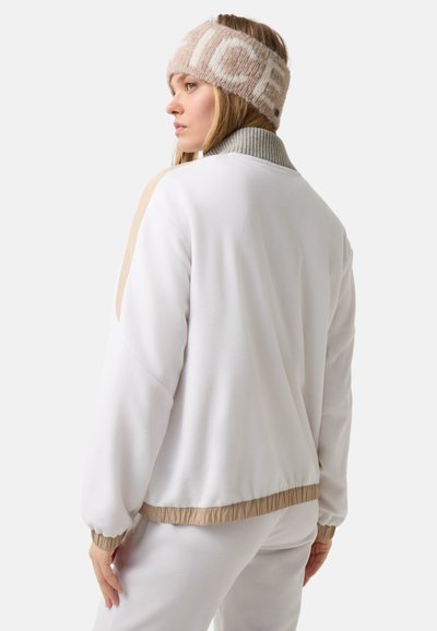 Bílá fleecová bunda s šedým žebrovaným límcem a béžovými detaily, s volným střihem a jemnou texturou. Skvěle doladěná s vzorovaným čelenkou.