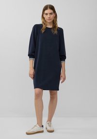 Marineblaues Sweatkleid mit lockerer Passform, langärmlig, mit weißen Streifen an den Bündchen und Rundhalsausschnitt. Getragen mit weißen Sportschuhen.