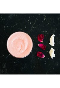 Scrub corpo rosa in una ciotola di vetro trasparente, circondata da petali di rosa rossa e pezzi di burro di karité bianco. Texture liscia, superficie lucida.