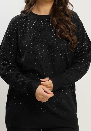 Femme portant un pull noir à manches longues avec de petits pois blancs, mains jointes et cheveux bruns bouclés sur l'épaule.