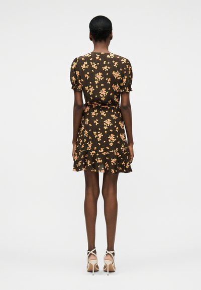 Γυναίκα με σκούρο δέρμα που φοράει καφέ κοντό φόρεμα με λουλούδια και βολάν στην τελειώμα, καθώς και μπεζ ψηλοτάκουνα σανδάλια, στέκεται γυρισμένη πλάτη σε λευκό φόντο.