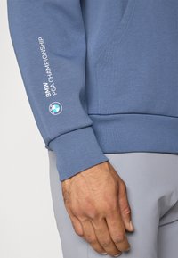 Sweatshirt azul com punhos canelados. A manga esquerda apresenta o texto "BMW PGA CHAMPIONSHIP" em branco e um logótipo circular da BMW.