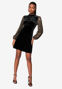 Fekete velúr mini ruha texturált arany mintás hosszú ujjakkal, kerek nyakkivágással és szabott derékkal, fekete, pántos magassarkúkkal párosítva.