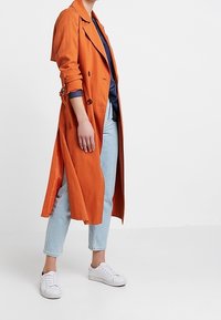 Lungo trench coat arancione realizzato in tessuto leggero, con un design doppiopetto e maniche arrotolate, abbinato a jeans di denim chiaro e scarpe da ginnastica bianche.
