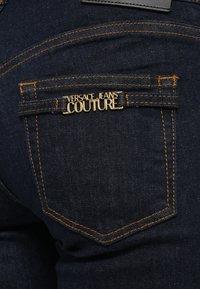 Sötétkék farmernadrág hátulján egy zseb található, amelyen a "VERSACE JEANS COUTURE" arany logó látható, kiemelve a kontrasztos sárga varrással.
