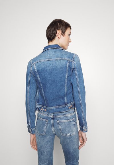 Personne avec les cheveux courts de dos, portant une veste en denim bleu ajustée et un jean denim bleu assorti, devant un fond uni.