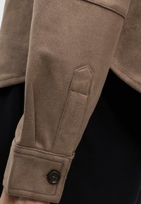 Manche de veste en daim marron avec détail de bride cousue et poignet noir boutonné sur fond noir.