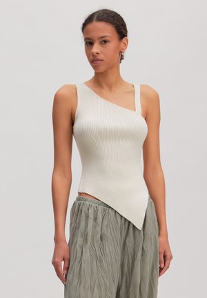 Jonge vrouw die een mouwloze, asymmetrische witte geribde top en losse, gekreukelde grijs-groene broek draagt, staand tegen een effen achtergrond.