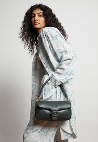 Vihreä nahkainen käsilaukku kultasävyisellä soljella, suorakaiteen muotoinen ja lyhyellä kahvalla, yhdistetty vaaleansinisellä marmorioidulla paitamekolla.