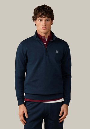 Jonge man die een marineblauwe trui met een kwart-ritssluiting draagt over een bordeaux poloshirt, staand tegen een effen beige achtergrond.