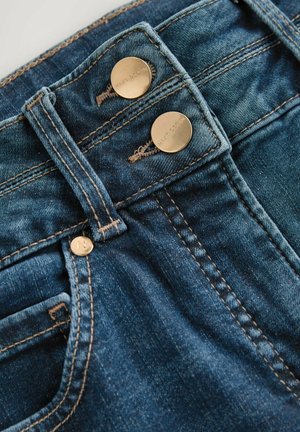 Detalii ale blugilor din denim, prezentând două butoane aurii, material albastru închis, cusături contrastante, fermoar frontal și o mică buzunar pe lateral.