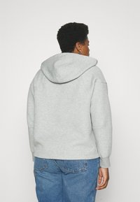 Gai pelēks džemperis ar lielu, noapaļotu kapuci, ribotām piedurknēm un brīvu piegriezumu, kombinēts ar ziliem džinsiem, skatīts no aizmugures.