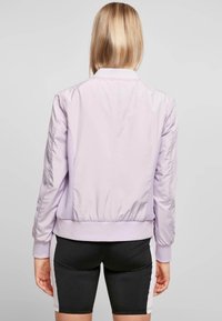 Jasnofioletowa kurtka bomberka o gładkiej, błyszczącej fakturze, prążkowanych mankietach i dolnym wykończeniu oraz siateczkowych panelach bocznych. Czarne szorty z białymi akcentami.