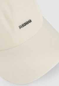 Cremefarvet kasket med en glat tekstur, der har et sølvfarvet "REISS" logo foran og en let buet skygge.