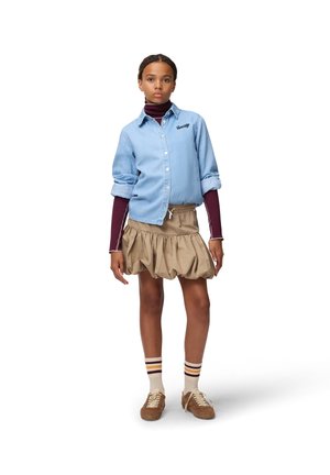 Persona joven de pie, con camisa azul claro abotonada sobre un jersey de cuello alto burdeos, falda beige fruncida, calcetines a rayas y zapatillas marrones.
