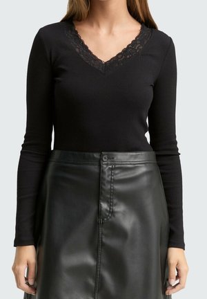 Femme portant un haut noir à manches longues avec col en V orné de dentelle et une jupe en cuir noire taille haute avec fermeture par bouton et fermeture éclair.