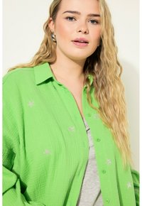 Zelená košile na knoflíky s texturovanou tkaninou, zdobená jemným vyšitím stříbrných hvězd. Nosená přes šedý top, s uvolněným střihem.