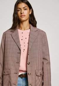 Blazer a cuadros en tonos rosa y gris, con un cuello con muesca, botones, dos bolsillos, superpuesto a una sudadera rosa con acentos metálicos.