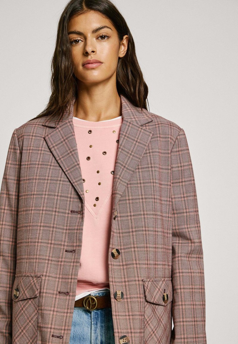Blazer a quadri nelle tonalità di rosa e grigio, con colletto a rever, bottoni, due tasche, indossato sopra un maglione rosa con dettagli metallici.