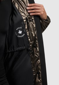 Fekete külső kabát fényes aranyszínű belsővel. Logó, patentgombok és húzózsinór jellemzi. A texturált anyag sima és tükröződő.