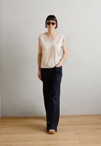 Lichtbeige V-hals t-shirt, losse fit, gecombineerd met donkerblauwe wijde jeans. Model draagt grote zonnebrillen en houten klompen op een houten vloer.