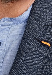 Marinblå kavaj med snedställda slag, texturerad tyg och en orange accent på slaget. Ljusblå skjorta med krage under.