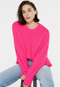 Helles pinkes gestricktes Sweater mit Rundhalsausschnitt, lockerer Passform und gerippten Bündchen, kombiniert mit hellblauen Jeans. Weiche Textur, lässiger Stil.