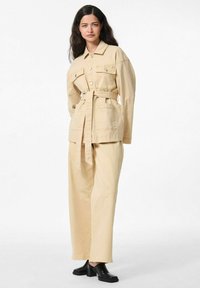 Veste en coton beige avec poches avant et ceinture, associée à un pantalon large beige et des mules à talons bloc noires. Texture lisse et coupe ajustée.
