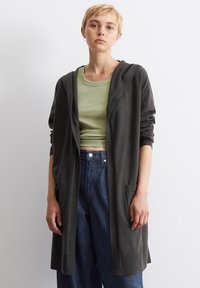 Mørkegrå lang cardigan med hette, med frontlommer og ribbestrikkede mansjetter, parret med en lys grønn tettsittende topp og high-waisted denimjeans.
