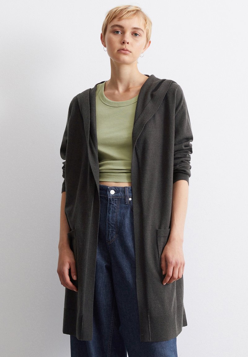 Mørkegrå lang cardigan med hette, med frontlommer og ribbestrikkede mansjetter, parret med en lys grønn tettsittende topp og high-waisted denimjeans.